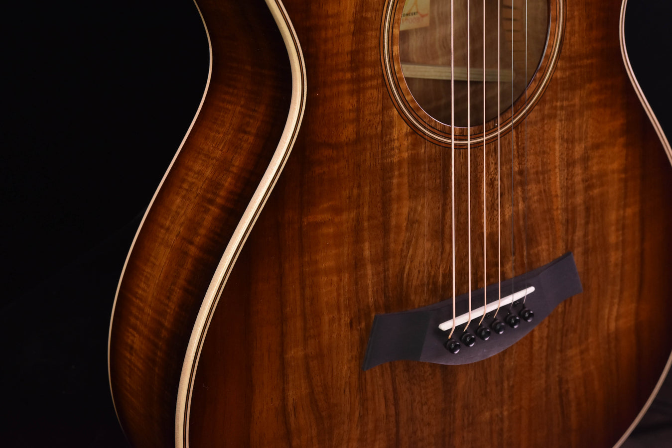 Koa acoustic guitar showing bridge area, dark bridge plate, and six tuning posts