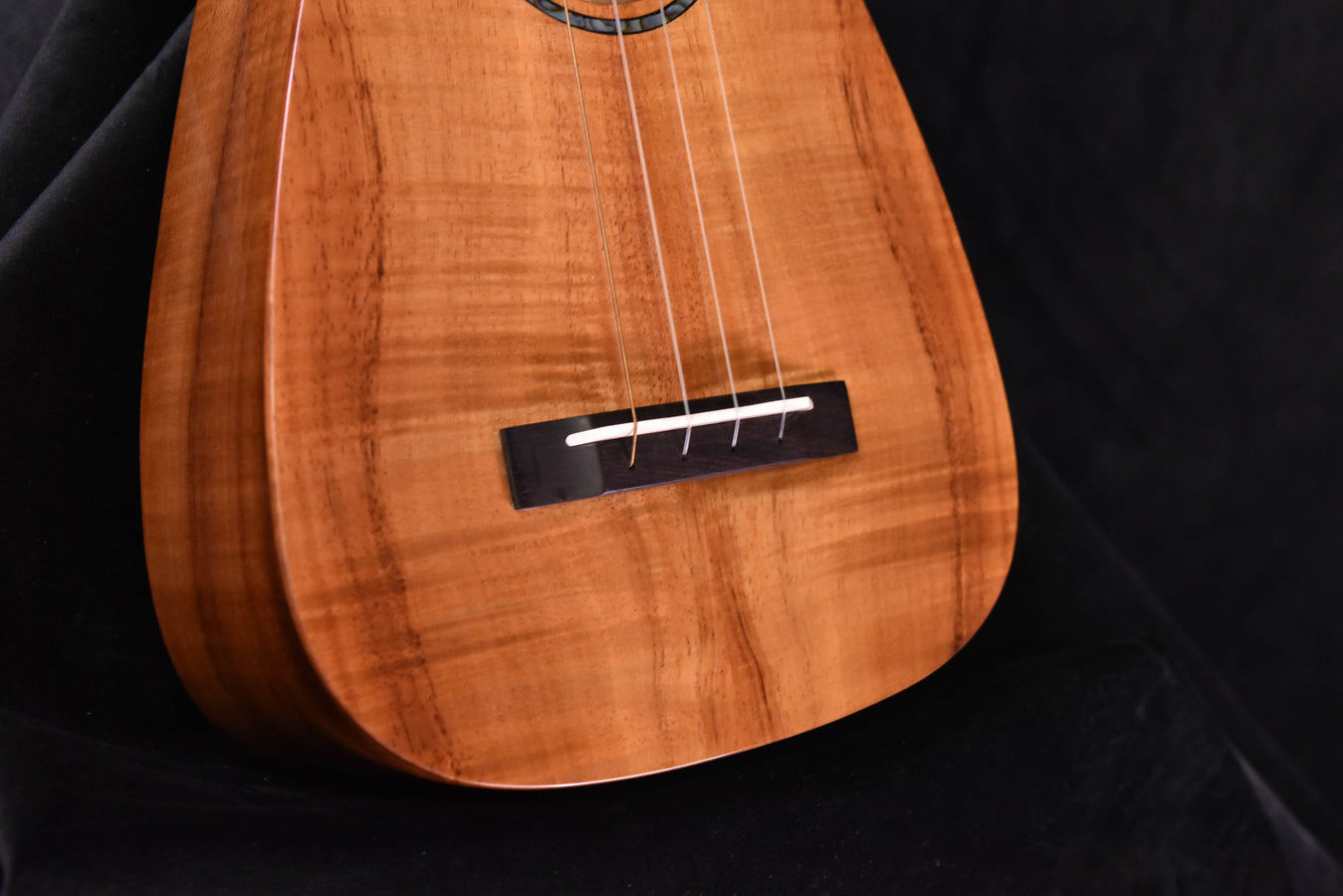 Koa tenor ukulele lower bout with black bridge and visible wood grain